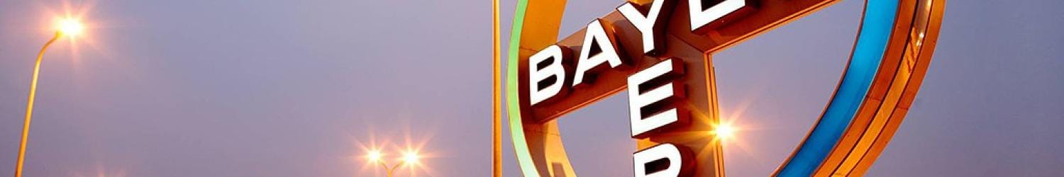 Illuminated Bayer logo on a large signpost with buildings and streetlights in the background during dusk.