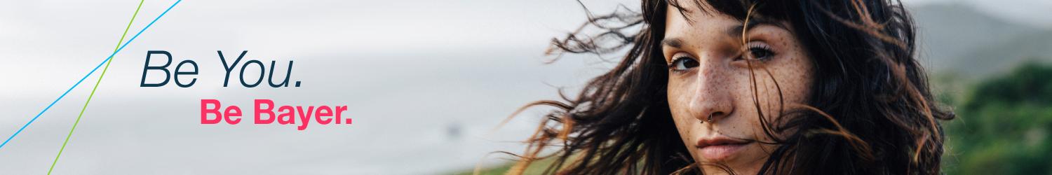 Person with long hair looking at the camera with greenery in the background. Text reads, "Be You. Be Bayer.