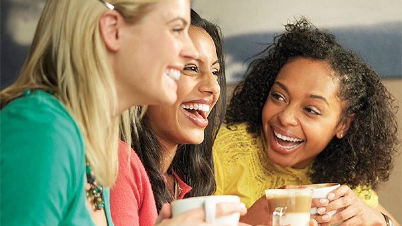 Three women sitting close together, smiling and holding mugs, appear to be enjoying a conversation in a casual setting.