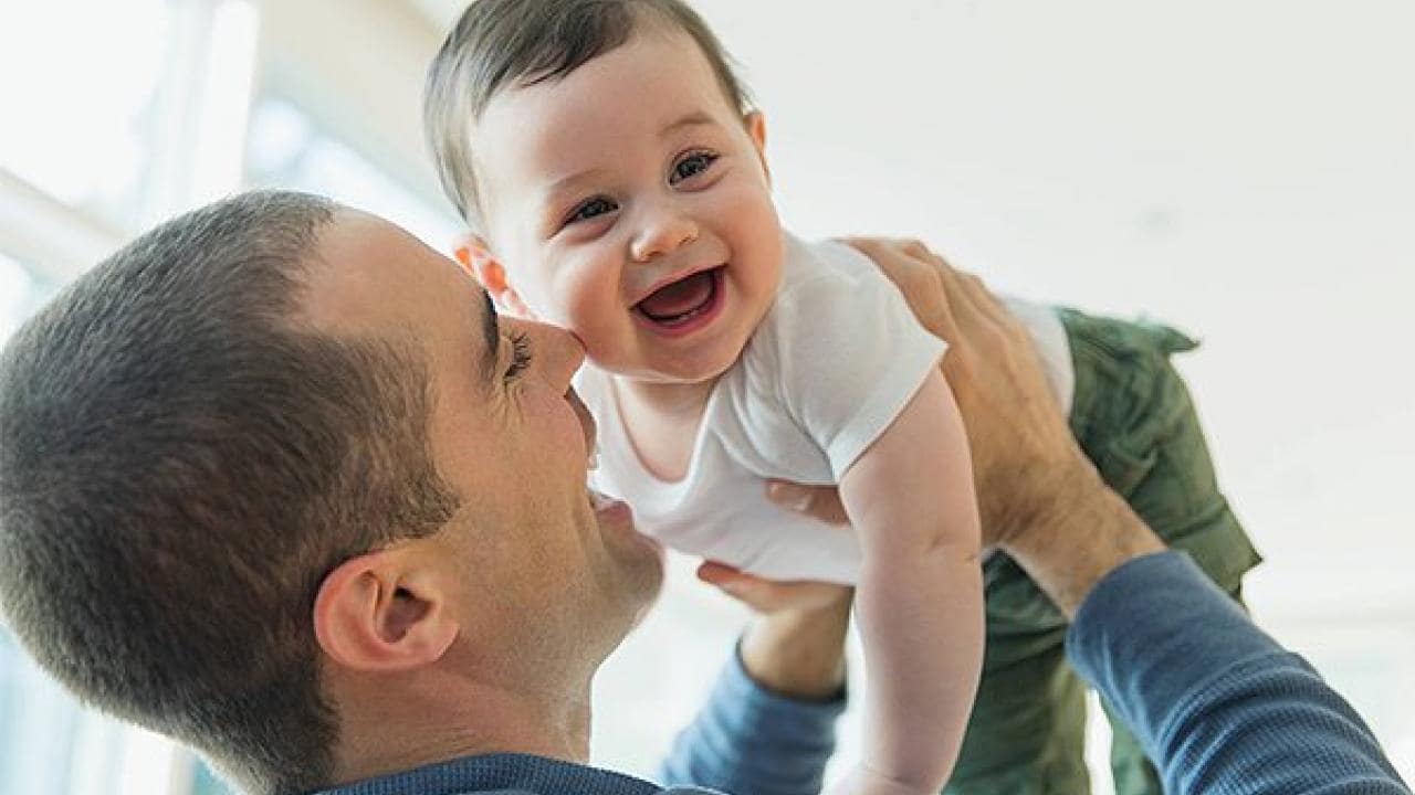 A smiling man holds a happy baby up in the air indoors, both wearing casual clothing.