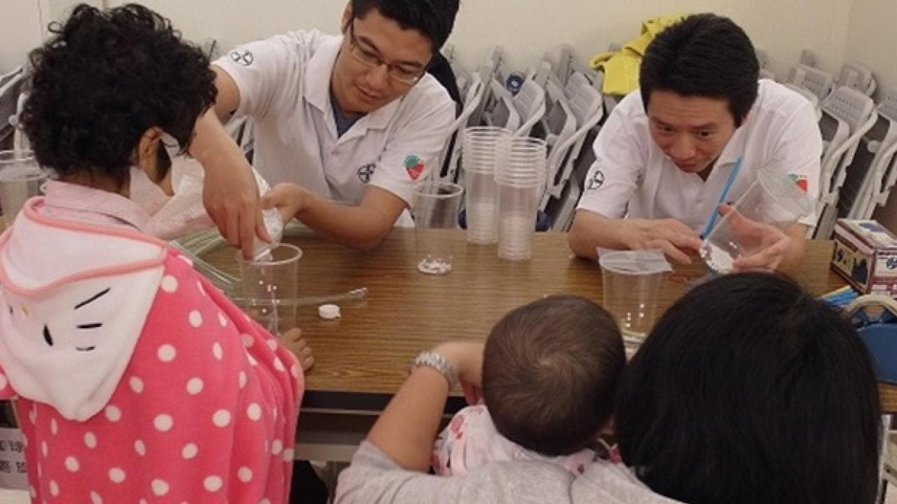 Two men conduct a science experiment at a table with transparent cups, while a woman with two children watches.