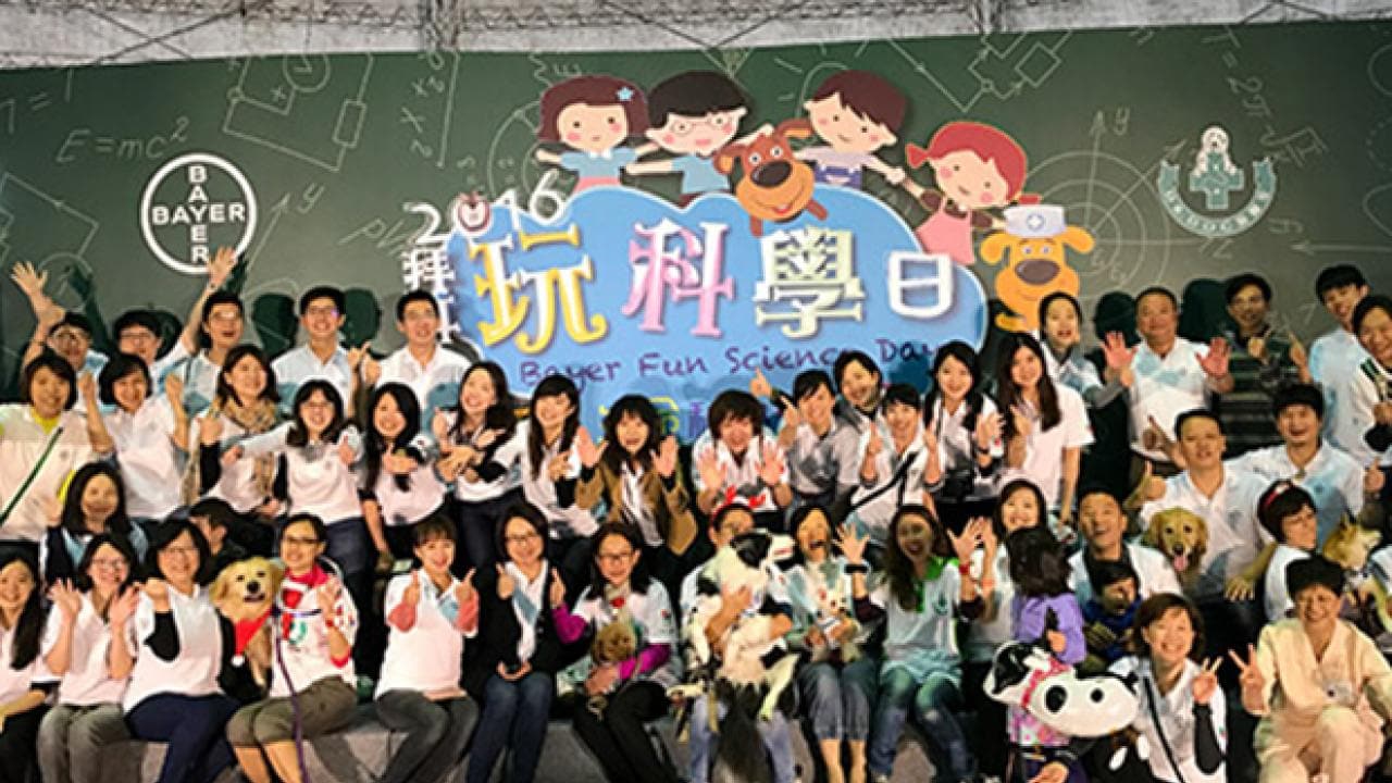 A large group of people pose together, some holding dogs, in front of a backdrop that reads "Bayer Fun Science Day 2016" with illustrated children and scientific symbols.