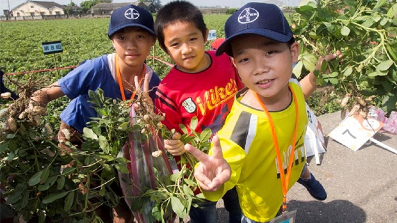 Three children holding plants and posing outdoors, wearing baseball caps and casual clothing, with fields and houses in the background.