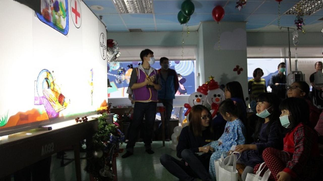 Children watching a puppet show in a decorated room. Some are wearing masks. There are balloons and snowmen decorations.