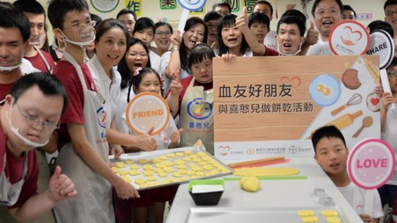 A group of people, some wearing aprons and face masks, gather around a table with cookie dough. They hold signs with words like "Friend" and "Love.