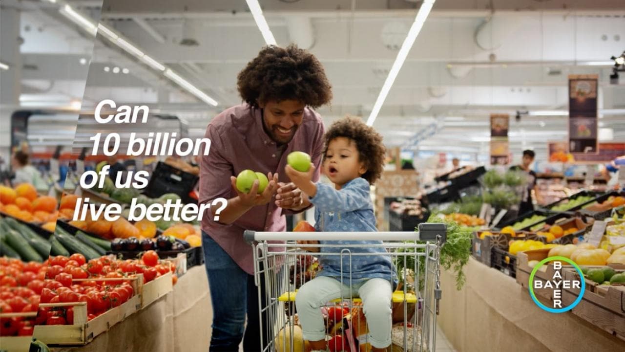 A person and a child shop for fruits in a grocery store. Nearby text reads, "Can 10 billion of us live better?" and a Bayer logo is visible.