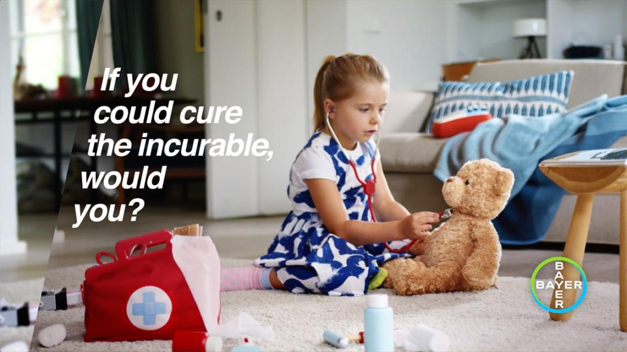 A young girl in a blue dress uses a stethoscope on a teddy bear. A red medical kit is nearby. Text reads, "If you could cure the incurable, would you?" Bayer logo is visible.