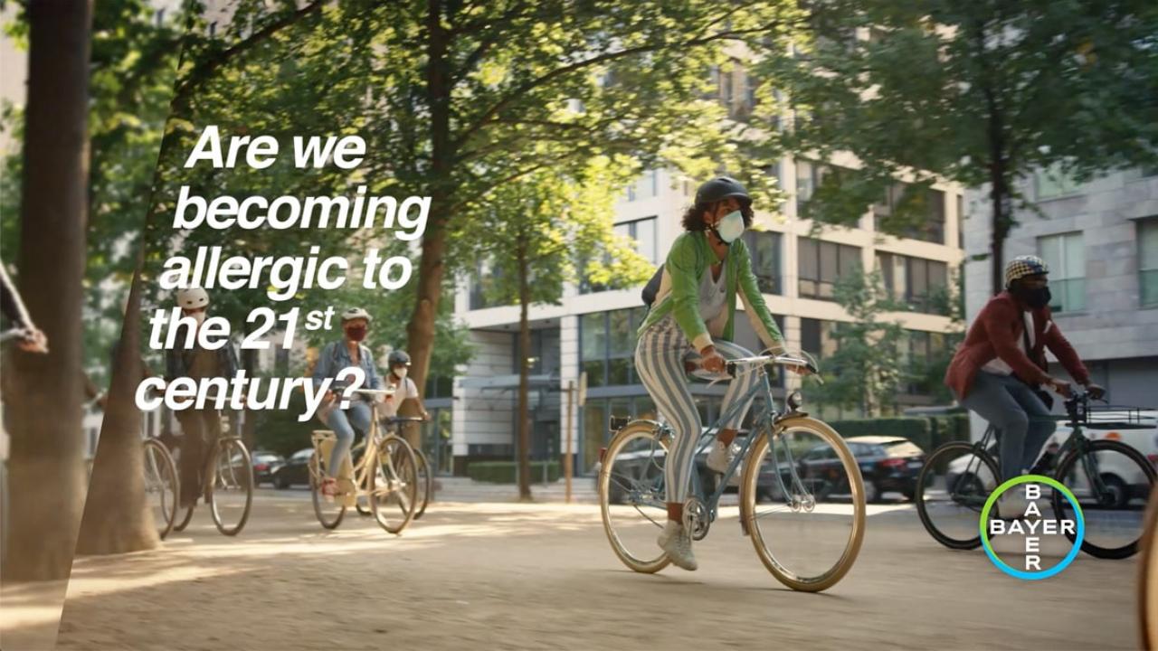 People riding bicycles in a city setting. Text on the image reads, "Are we becoming allergic to the 21st century?" with a Bayer logo in the corner.