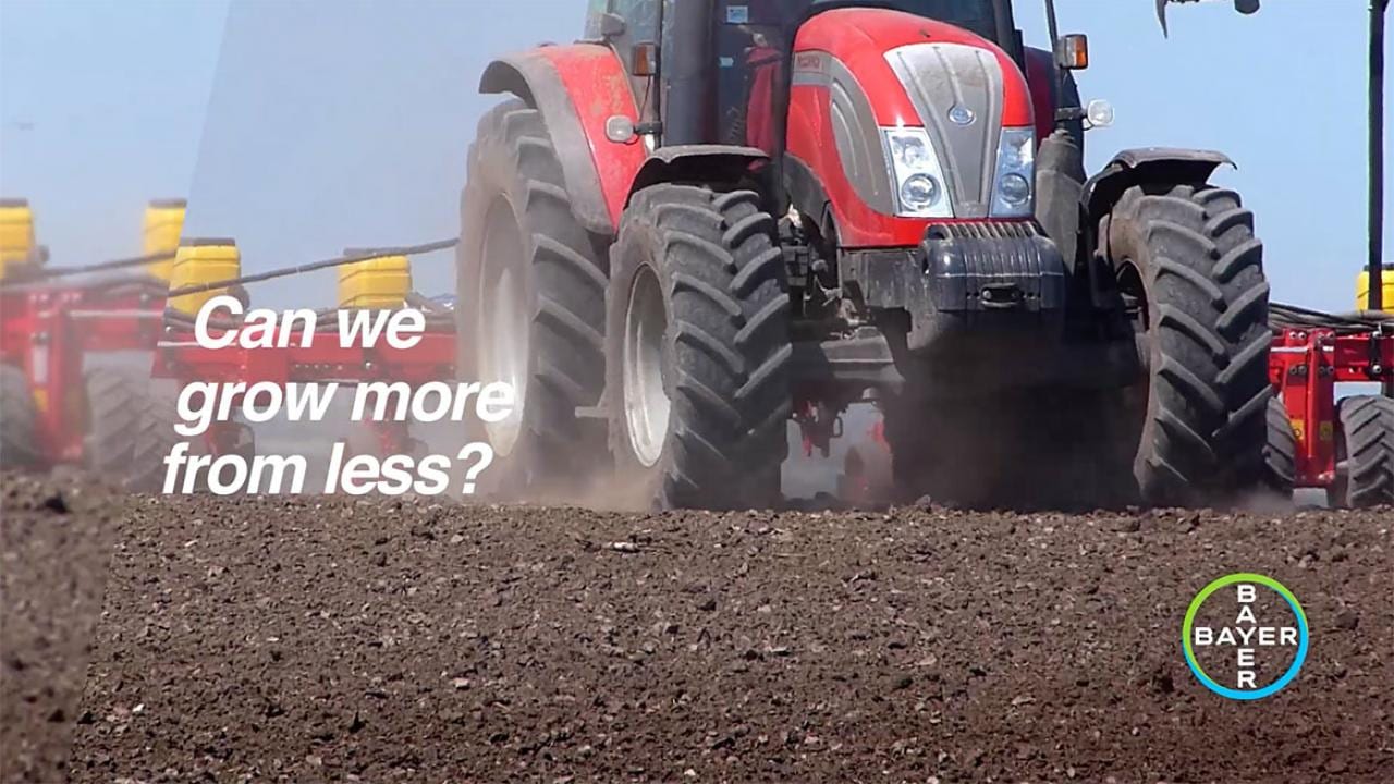 A red tractor plows a field with the text, "Can we grow more from less?" and a Bayer logo in the corner.