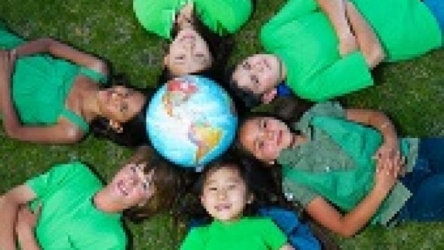 Six children in green shirts lie on grass, forming a circle around a globe placed in the center.