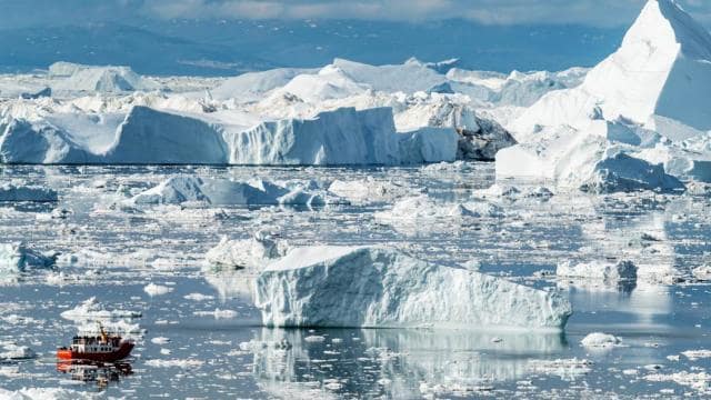 A small boat navigates through a sea filled with large icebergs and smaller ice chunks, set against a backdrop of a distant mountainous landscape.