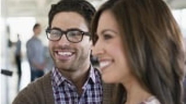 Two people in a well-lit room are smiling and looking in the same direction. The person on the left wears glasses and a plaid shirt, while the person on the right has long brown hair.