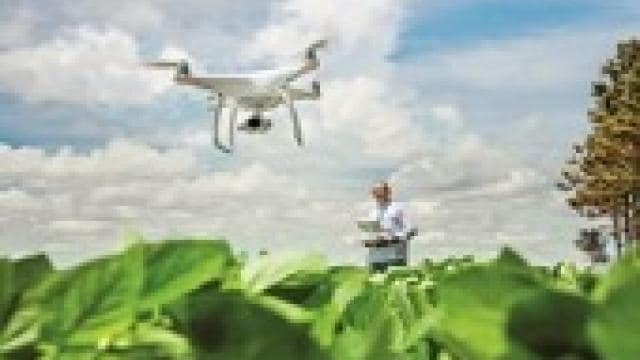 A person operates a drone over a lush green field, with scattered clouds in the blue sky.
