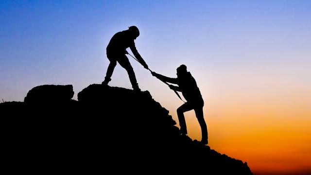Two silhouetted figures on a rocky slope, one helping the other up, against a backdrop of a colorful sunset sky.