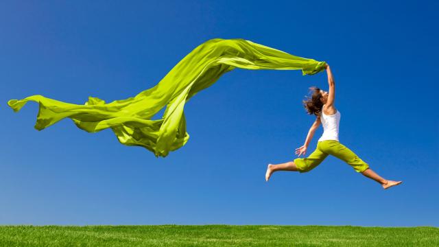 A person in green pants and a white shirt leaps across grass, holding a flowing green fabric, set against a clear blue sky.