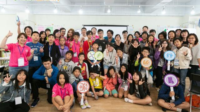 A diverse group of people, including adults and children, pose in a room. Some hold circular signs with various symbols. A green and yellow logo is visible in the top left corner.