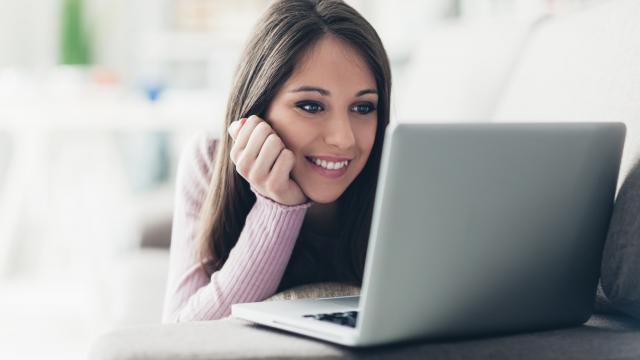 A person with long hair lying on a couch looks at a laptop with a smile.