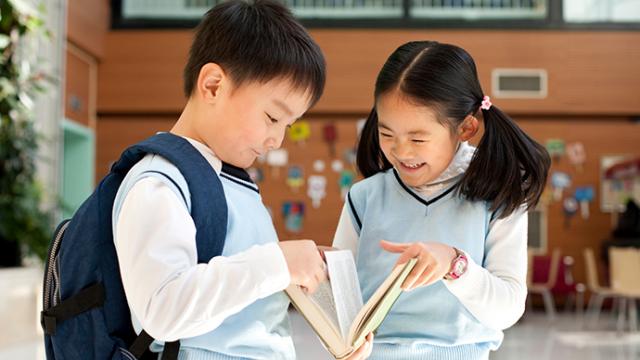 Two children in school uniforms read a book together indoors. One child wears a backpack, and both are smiling.