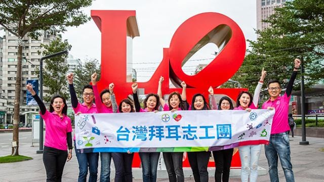 A group of people in pink shirts hold a banner in front of a large "LOVE" sculpture outdoors.