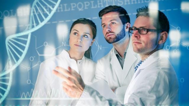 Three researchers in lab coats examine a translucent DNA strand and molecular structures on a digital screen.