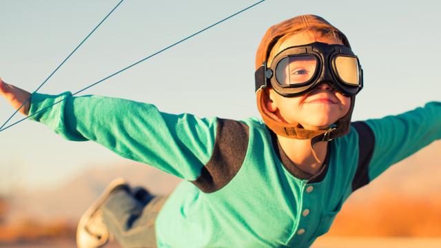 Child wearing aviator goggles and cap, arms outstretched, appears to be flying against a blurred outdoor background.