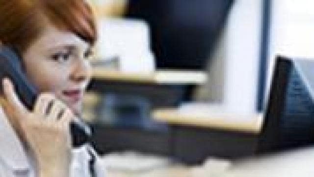 A person with red hair is sitting and talking on a landline phone while looking at a computer screen.