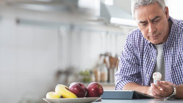 A man in a checkered shirt looks at a pill bottle in a kitchen, with a fruit bowl and glass of water on the table.