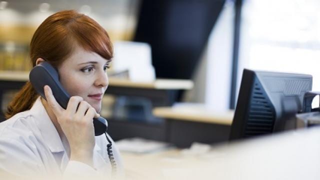 A woman with red hair is sitting at a desk, talking on a landline phone, and looking at a computer monitor.