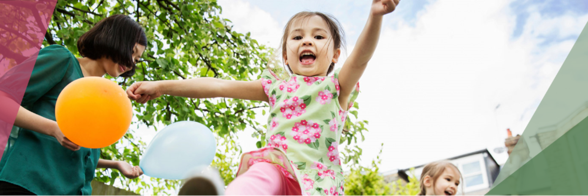 A joyful child jumps with arms outstretched, holding balloons, accompanied by an adult and another child, with a leafy tree and blue sky in the background.