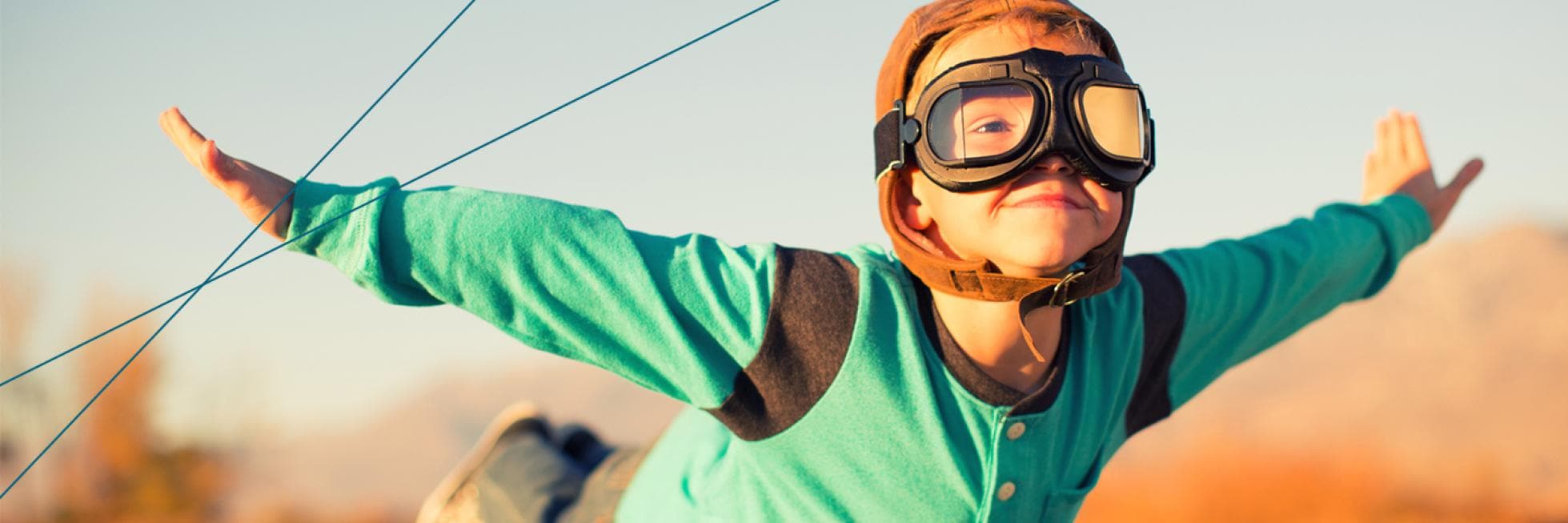 Child wearing aviator goggles and cap, arms outstretched, appears to be flying against a blurred outdoor background.