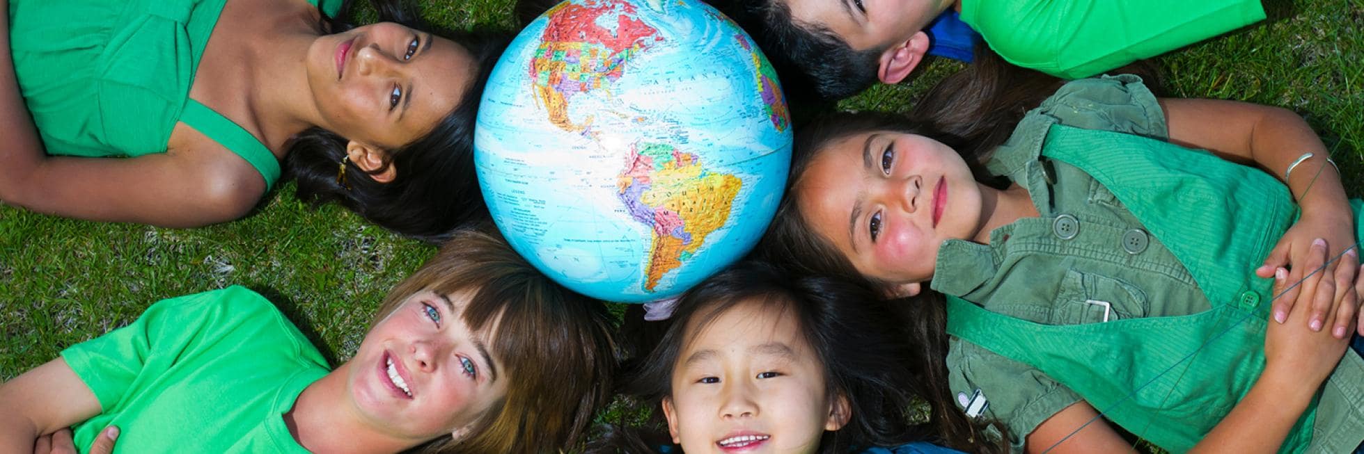 Five children lie on grass in a circle, heads together, surrounding a globe. They wear green shirts and smile at the camera.